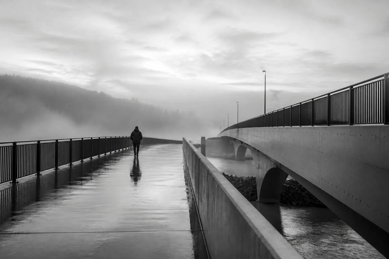 Black-and-white photograph of an anonymous figure crossing a rain-darkened bridge in mist